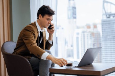 Telefon görüşmeleri sırasında dizüstü bilgisayarla çalışan profesyonel iş adamının portresi. Akıllı yönetici dizüstü bilgisayar ve kahve fincanı ile veri analizi yaparak arama yapıyor. Gökdelen manzaralı. Süslü.