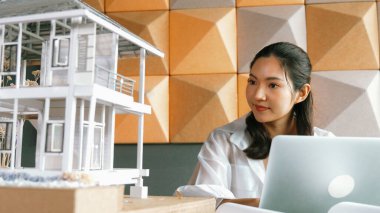 Genç ve güzel mimar mühendisin portresi çatı malzemelerini kontrol ediyor ve ofisteki masaya yerleştirilmiş proje planı ve ev modeliyle dizüstü bilgisayar kullanıyor. Yaratıcı iş tasarımı konsepti. Yönlendirici.