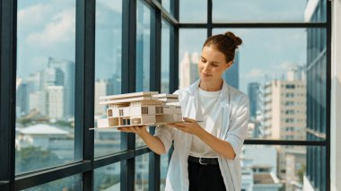 Genç, beyaz, güzel mimar mühendisi ev modelini yükseltirken şehir gökdelen manzarası dikkatlice inceleniyor. İnşaat mühendisliği, mimari stüdyosu. Proje planlama. İzleme