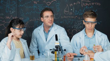 Yaratıcı öğretmen masadaki liseli çocukla mikroskop ve renkli sıvıyla test tüpüyle konuşurken kimyayı tahtaya doğrultuyor. BTMM sınıfında laboratuvar önlüğü giyen genç bir öğrenci. Öğretim.