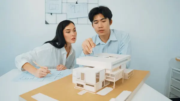 Closeup of skilled architecture engineer team measure and decide house model while working together and discussing about house structure at meeting room with blueprint placed behind. Immaculate.