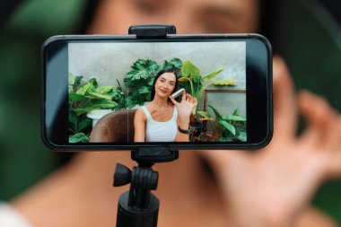 Arkadan bakan kadın, telefon ekranında yeşil bitki bahçesi içeriği ekranında doğal güzellik ve kozmetik dersi veriyor. Güzellik blogcusu sosyal medyum izleyicilere güzellik bakımını gösteriyor. Blithe