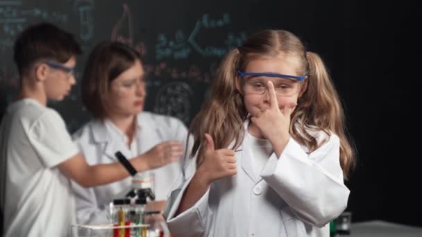 聪明的高加索女孩在老师和学生后面做实验时展示了她的大拇指 孩子们看着相机 而男孩们则在显微镜下看着黑板 写下了化学理论 Erudition — 图库视频影像