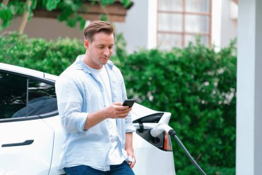 Modern çevre dostu adam EV şarj istasyonundan elektrikli aracı şarj ediyor. Evde şarj edilen pilleri optimize etmek için enerji kullanımını takip etmek için yenilikçi EV teknolojisi kullanımı. Eşzamanlama