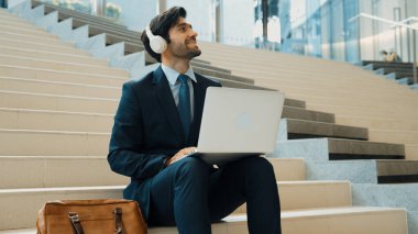 Profesyonel bir iş adamı dizüstü bilgisayarla çalışırken merdivenlerde oturuyor. Yetenekli proje yöneticisi kulaklıktan müzik dinliyor ve e-postaları kontrol ediyor ve pazarlama planı hakkında tartışıyorlar. Açık havada. Sevinçli..