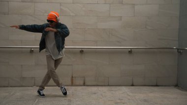 Asyalı akıllı hipster, kulaklık ve şık kıyafetler giyip hip hop şarkıları dinlerken gri arka planlı enerjik break dans gösterisi yapıyor. Açık hava sporu 2024. Hip hop hareketi. Yumuşak bir şekilde..