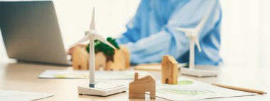 Temiz enerji kullanılarak temsil edilen yel değirmeni modeli ve temsil edilen ahşap blok dizüstü bilgisayarla çalışan iş adamının önünde masaya saçıldı. Geçmişi bulanıklaştırıyor. Tanımlama.
