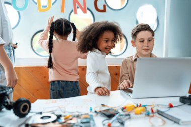 Sınıftaki çocuklar BTMM sınıfında mühendislik öğreniyor. Okul çocuğu sandalyede oturuyor ve uzaktan kumandalı arabayı dizüstü bilgisayarla kodluyor. Öğretmen ve diğer çocuklar etrafında. Not defteri, elektrik kablosu. Etkinlik.