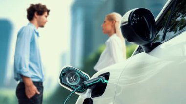 İş adamları otoparkta şarj olan elektrikli arabayı şarj ediyor. Akıllı akü durumu hologramı gösteriyor. Modern şehir yaşam tarzı ve karbondioksiti azaltmak için çevre dostu elektrikli araba kullanımı. İnceleyin