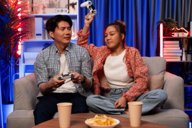 Mutlu çiftin oturduğu kanepe joysticklerle video oyunu oynuyor, meydan okuyan gülümseyen kadın, evdeki modern neon ışıklı oturma odasındaki mücadeleyi kazanmak için yumruk kaldırıyor. İnfobahn.