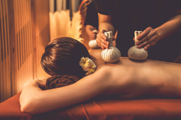 Hot herbal ball spa massage body treatment, masseur gently compresses herb bag on couple customer body. Serenity of aromatherapy recreation in warm lighting of candles at spa salon. Quiescent