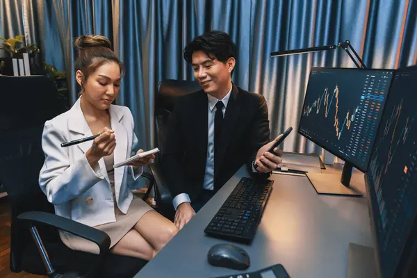 アジアのビジネスマンは 夜間のネオンライトでコンピュータ画面上の金融デジタル投資データベースを比較して ペンを指すパートナーに携帯電話の株式市場アプリケーションを示しています インフォバーン — ストック写真