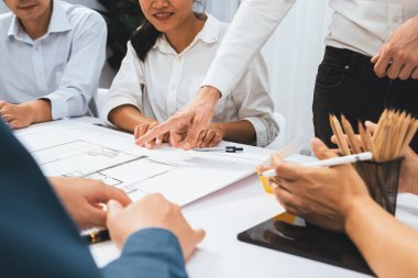 Mimari proje üzerinde birlikte çalışan çeşitli inşaat mühendisi ve müvekkil grupları, inşaat planını gözden geçirip toplantı masasında plan oluşturuyorlar. Tedbirli