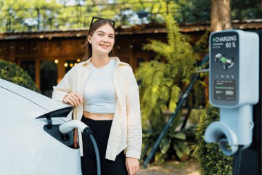 İlkbahar bahçesindeki açık kafede çalışan EV elektrikli araba şarjı, yeşil şehir sürdürülebilirliği ve çevre dostu EV otomobili için genç bir kadınla sürdürülebilir şehir seyahati elektrik ödüyor. Yararlı
