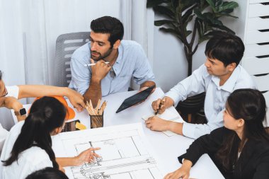 Mimari proje üzerinde birlikte çalışan çeşitli inşaat mühendisi ve müvekkil grupları, inşaat planını gözden geçirip toplantı masasında plan oluşturuyorlar. Tedbirli