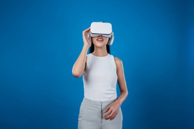 Mavi arka planı olan zeki bir kadın metaevrene bağlı VR kulaklık takıyor, gelecekçi siber uzay topluluğu teknolojisi. Zarif bir kadın sanal bir manzara gördüğü için heyecanlı. Halüsinasyon.