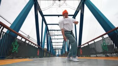 Profesyonel break dansçı Briç 'te sokak dansı yapıyor. Asyalı hippi dondurucu poz verirken kulaklık takıyor. Break dansçı, serbest stil sokak dansçısı konsepti. Açık hava sporu 2024. Yumuşak bir şekilde..
