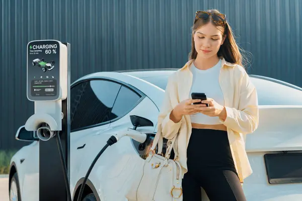 ショッピングバッグを持っている若い女性とスマートフォンを使用して 都市モール駐車場の充電ステーションからEvカーバッテリーを充電するために電気を支払います 現代の女性はエコカーで買い物をする エクスペディエント — ストック写真