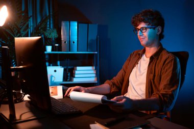 Genç işadamı, Neon Light Modern Ofis 'teki yeni proje surround çalışma masası için kağıt üzerinde tasarım düşünüyor. E-posta ile hediye göndermeden önce not defterine yeni bir iş düşünüyorum. Toplayıcı.