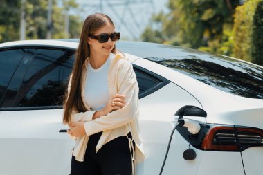 Genç bir kadın yeşil şehir parkındaki elektrikli aracını şarj ediyor. Batarya şarj istasyonu olan çevre dostu EV araba için kentsel sürdürülebilirlik yaşam tarzı. Yararlı