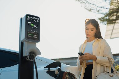 Alışveriş çantası tutan genç bir kadın, alışveriş merkezinin otoparkındaki şarj istasyonundan EV 'nin aküsünü şarj etmek için akıllı telefon kullanıyor. Modern kadın eco arabasıyla alışverişe gider. Yararlı