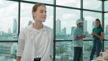 Genç ve güzel proje yöneticisi, mühendis ekibi ev tasarımı hakkında tartışırken kameraya poz veriyor. Modern ofiste ev modelinin yanında otururken kameraya bakan yetenekli bir iş kadını. İzleme