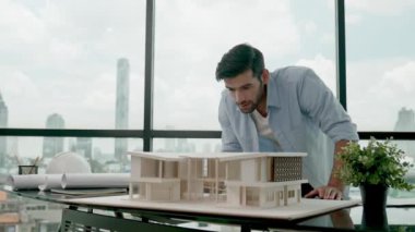 Yakışıklı beyaz mühendis veya proje yöneticisi ölçüm, teftiş, ev modeline ve bina planına bakarken şehir manzaralı panorama penceresinin yanında duruyor. İzleme