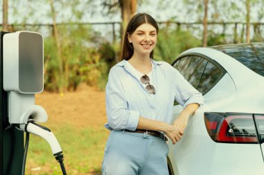 Genç bir kadın sonbahar gezisi sırasında sonbahar ormanı EV arabasında elektrikli araba için pilini şarj ediyor. Sonbahar tatilinde elektrikli araçla çevre dostu bir seyahat. Yükselt