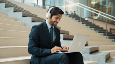 Kendine güveni tam bir iş adamı başarılı iş fikrini müzik dinlerken kutluyor. Happy yönetici projesi kulaklık ve takım elbise giyerken dizüstü bilgisayarla çalışıyor. Yatırımcı merdivenlerde oturuyor. Sevinçli..
