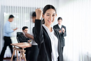 Kameraya bakan mutlu Asyalı genç iş kadını portresi, dinamik iş toplantısındaki iş adamları hareketinin hareket bulanık arka planıyla yumruğunu kaldırıyor. Habiliment
