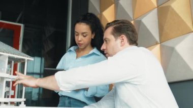 Proje yöneticisi bina tasarımı hakkında tartışırken ev modelini işaret ediyor. Mimar mühendis ekibi bina yapısını kontrol ederken iş arkadaşı tablet analiz verilerini kullanıyor. Takım çalışması. Yönlendirici.