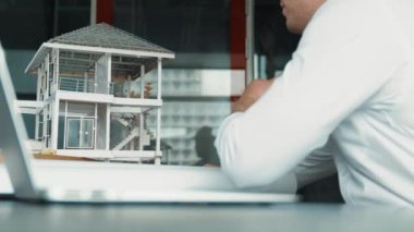 Kabiliyetli beyaz mimar el yapımı ev tasarımını dizüstü bilgisayardaki model ve plan ile karşılaştırın. Laptop analiz verilerini kullanan profesyonel proje yöneticisi. Eline odaklan. Yönlendirici.
