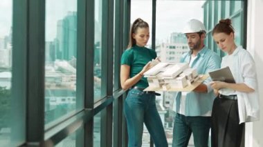 Proje yöneticisi ev modelini elinde tutarken, mimar mühendis takımı da ev tasarımı veya bina inşaatı hakkında fikir alışverişinde bulunuyor. Şehrin penceresinin yanında iç mimar duruyor. İzleme