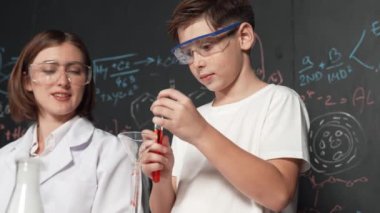 Akıllı çocuk, öğretmen STEM fen dersinde tavsiye verirken deney kabına kimyasal sıvı döker. Öğrenci renkli çözeltileri karıştırırken laboratuvarda tahtada duran yazılı kimya teorisi. Etkinlik.