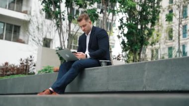 Yetenekli erkek lider Green City 'deki yatırım verilerini analiz ederken aynı zamanda dizüstü bilgisayarını kullanarak telefon gönderme raporu üzerinde çalışıyor. Parkta ya da şehir merkezinde otururken yatırımcı görünümlü mutlu pazarlama planı. Urbane.