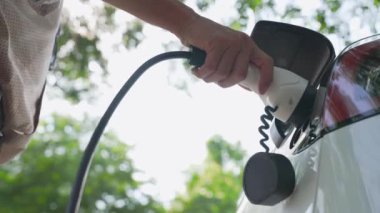 Genç adam, yaz mevsiminde açık yeşil yay yeşilliklerindeki şarj istasyonundan elektrikli araba aküsünü şarj etmek için EV şarj aleti koymuş. Yeniden şarj edilebilir elektrikli araç için sürdürülebilir enerji
