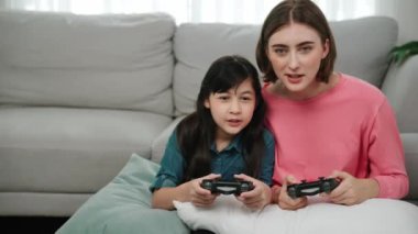 Mutlu anne ve enerjik kız oyun oynuyor ve birlikte vakit geçiriyorlar. Kafkasyalı anne, elinde joystick tutarken çocuğunun yanında oturuyor ve rahat oturma odasında kızıyla oyun oynamaya odaklanıyor. Pedagoji.
