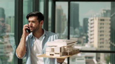 Gülümseyen Kafkasyalı mühendis ev modelini sağa sola çeviriyor ve iş arkadaşına telefon ederken hata noktasını kontrol ediyor. Profesyonel yönetici teftiş, kontrol, mimari modele bakmak. İzleme