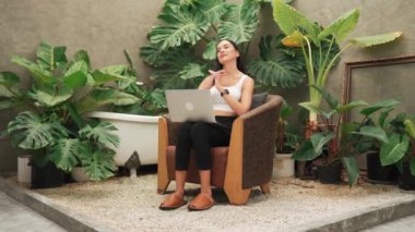 Uzaktan çalıştıktan sonra vücudunu esneten modern genç kadın ya da tropikal bitkilerle birlikte minimalist beton mimari stil yaz egzotik bitki yetiştirme bahçesinde yalnız başına dinleniyor. Blithe