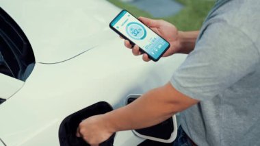 Modern yaşam tarzı adamı EV şarj cihazını ev şarj istasyonuna yerleştirir ve akıllı telefondan EV batarya durumunu etkinleştirir. Gelecekçi enerji sürdürülebilirliği ve elektrikli araba. İnceleyin