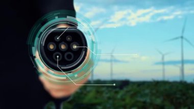 Kameranın önüne odaklanmış EV şarj cihazı fütüristik akıllı dijital EV batarya durum hologramını gösteriyor. EV araba şarjı, ekolojik sürdürülebilir güç için rüzgar türbininin yenilenebilir enerjisini kullanıyor. İnceleyin