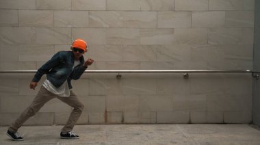 Asyalı akıllı hipster, kulaklık ve şık kıyafetler giyip hip hop şarkıları dinlerken gri arka planlı enerjik break dans gösterisi yapıyor. Açık hava sporu 2024. Hip hop hareketi. Yumuşak bir şekilde..