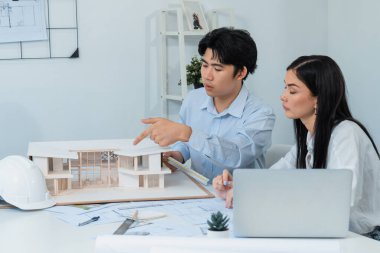 Profesyonel Asyalı erkek mimar ev modelinin uzunluğunu ölçmek için cetvel kullanırken, beyaz tenli genç meslektaşı ev modeliyle toplantı masasındaki verileri analiz etmek için dizüstü bilgisayar kullanıyor. Tertemiz..