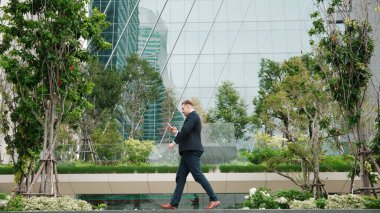 Kafkasyalı işadamının akıllı telefona bakması ve yeşil eko şehrindeki modern binaya yürümesi. Yetenekli lider telefon kullanarak raporları kontrol ederken işyerine gidiyor. Yaşam tarzı. Urbane.