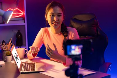 Smiling creative young beautiful Asian influencer recording on camera with positive podcast speech at night time, using laptop for presentation online at pink neon light modern studio. Stratagem. clipart