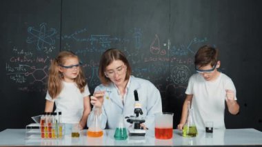 Öğretmen tavsiye verirken beyaz çocuk kimyasal sıvıyı karıştırıyor. Profesyonel eğitmen labaratuar kıyafeti giymiş, renkli solüsyonla dolu deney kabıyla sofrada çeşitli öğrencileri arıyor. Etkinlik.