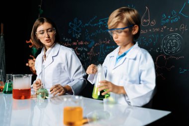 Labaratuvarda yardımcı öğretmen öğrenci laboratuvar önlüğü ve gözlük takıyor ve STEM sınıfında kimya bilimi üzerine deney yapıyorlar. Öğrenci derneği, beher içinde sarı sıvıya indi. Etkinlik.