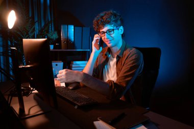 Genç iş adamı akıllı telefondan müşteriyi ya da iş arkadaşını arıyor, neon modern ofisteki yaratıcı sunum surround çalışma masasının bilgisayar ekranına bakıyor. Yeni proje kavramı. Toplayıcı.