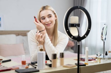 Sosyal medya için güzellik ve kozmetik video içeriği hazırlayan genç bir kadın. Seyirciye veya takipçiye nasıl sıvı ruj sürüleceğini gösterirken kamera ve ışık halkası kullanan bir güzellik blogcusu. Blithe