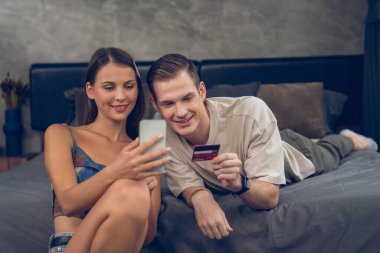 Genç çift, kredi kartıyla ödemek için internet üzerinden ödeme uygulaması ve dijital cüzdan kullanarak yatak odasında oturuyor. Mobil internet üzerinden alışveriş ve modern alımlar. Açılıyor